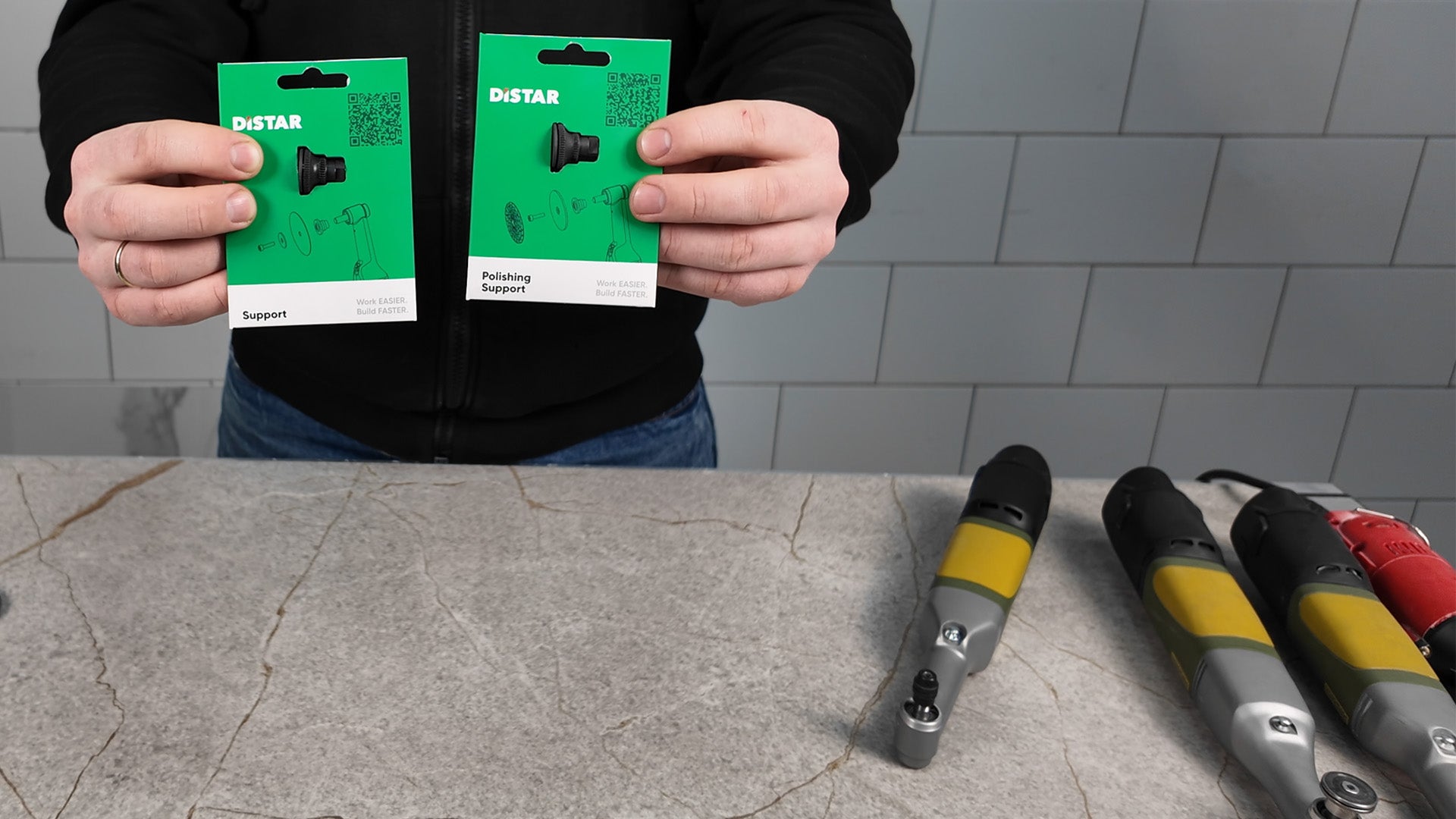 Person holding two green DISTAR packaging against a tiled wall background with tools on a counter.